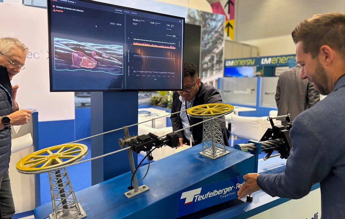 Photo of a conference floor, showing a mini simulation of ski lift, with a steel wire rope going around and a triple camera setup inspeccting the rope setup. 3 men are looking at the setup. Behind the rop is a TV displaying damages on the rope.